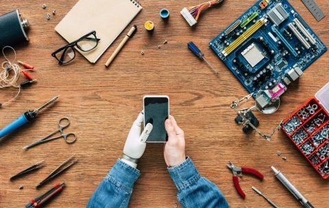 cropped image of  electronic engineer with prosthetic arm using smartphone at table surrounded by