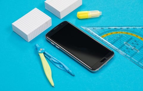 Two piles of white scratch paper, a pair of compasses, one ruler, a yellow text marker and black smartphone on azure background isolated
